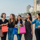 Sunny outdoor shopping street with diverse group of travelers carrying shopping bags, modern cityscape, vibrant atmosphere, India Tours & Travels.
