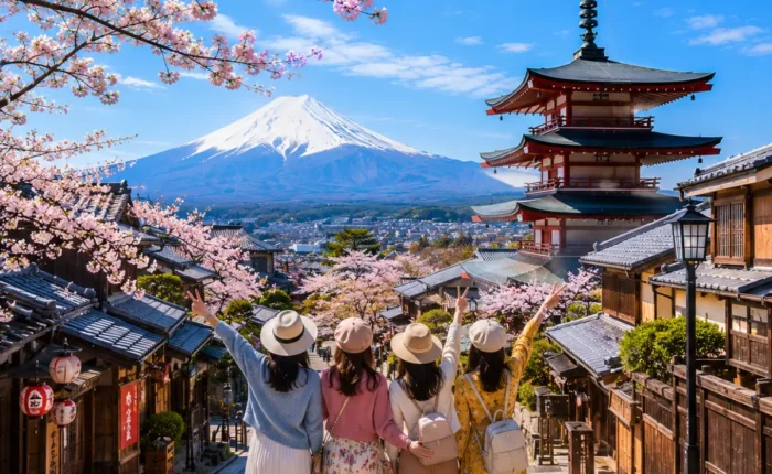 Women-only Japan tour featuring four female travellers exploring Kyoto's historic streets with the iconic Yasaka Pagoda, Mount Fuji, traditional wooden houses, and blooming cherry blossoms under a bright blue sky.