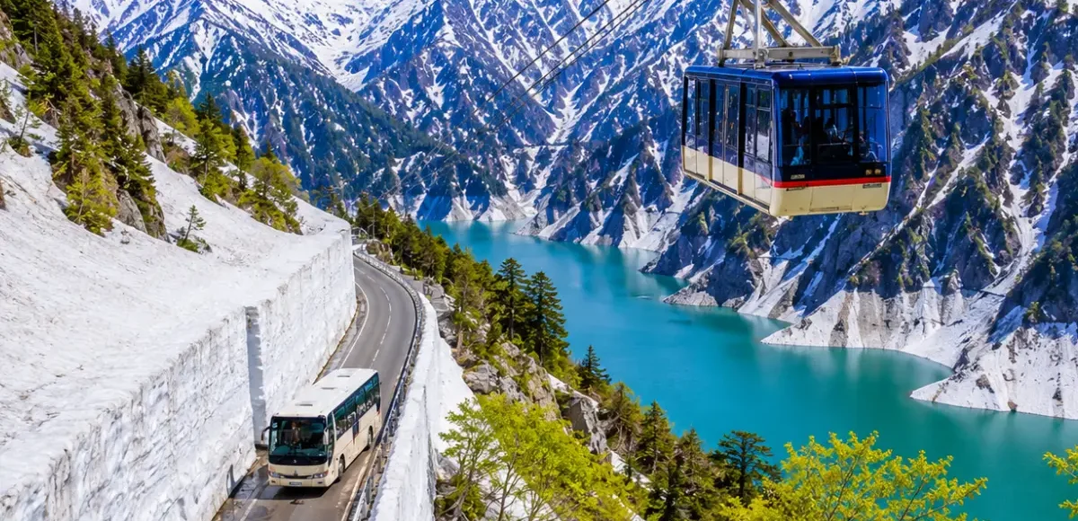 Luxury Japan Alpine Route featuring the iconic Tateyama Kurobe Snow Corridor, crystal-clear turquoise Kurobe Lake, panoramic snow-covered peaks, an alpine cable car, and a scenic mountain road under bright blue skies. No people.