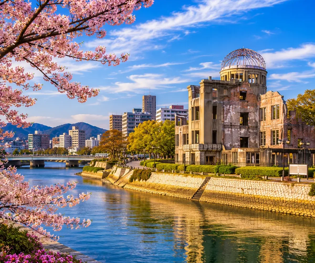 Luxury Hiroshima cityscape featuring the Hiroshima Peace Memorial (Atomic Bomb Dome), a tranquil riverside, blooming cherry blossom trees, modern skyline, and crystal-clear blue skies in bright daylight. No people.
