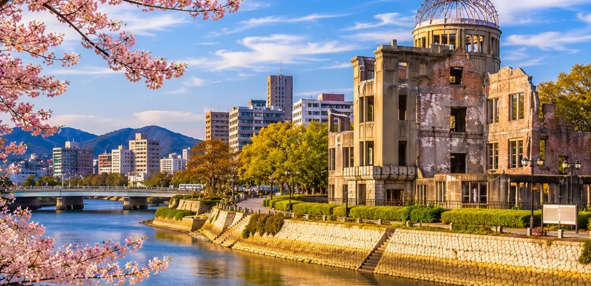 Luxury Hiroshima cityscape featuring the Hiroshima Peace Memorial (Atomic Bomb Dome), a tranquil riverside, blooming cherry blossom trees, modern skyline, and crystal-clear blue skies in bright daylight. No people.