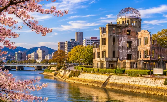 Luxury Hiroshima cityscape featuring the Hiroshima Peace Memorial (Atomic Bomb Dome), a tranquil riverside, blooming cherry blossom trees, modern skyline, and crystal-clear blue skies in bright daylight. No people.