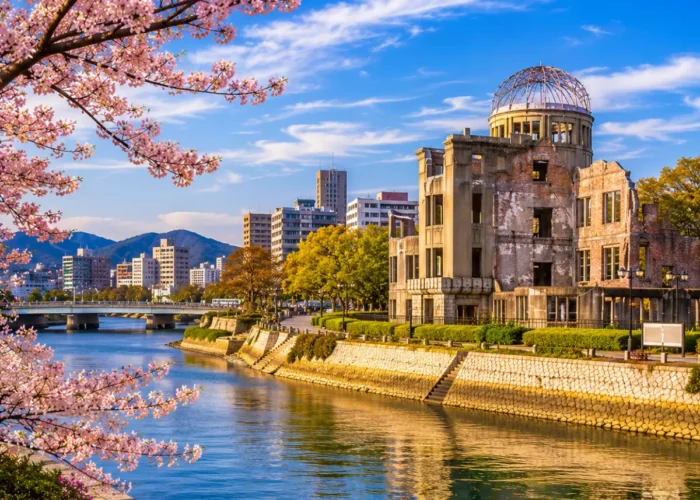 Luxury Hiroshima cityscape featuring the Hiroshima Peace Memorial (Atomic Bomb Dome), a tranquil riverside, blooming cherry blossom trees, modern skyline, and crystal-clear blue skies in bright daylight. No people.