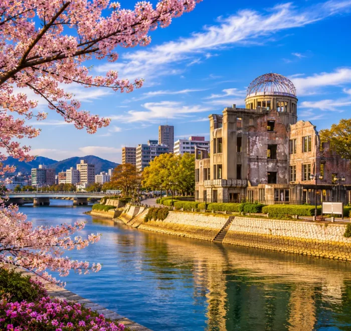 Luxury Hiroshima cityscape featuring the Hiroshima Peace Memorial (Atomic Bomb Dome), a tranquil riverside, blooming cherry blossom trees, modern skyline, and crystal-clear blue skies in bright daylight. No people.