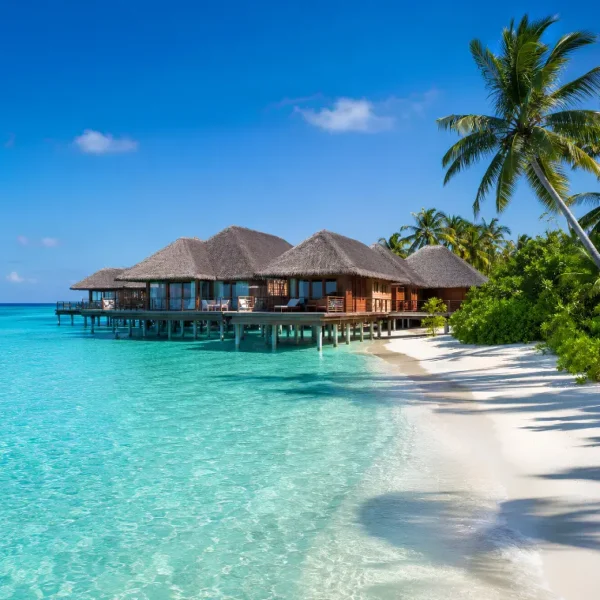 Maldives resort with overwater villas, turquoise lagoon, white sand beach, palm trees, and clear blue daytime sky.