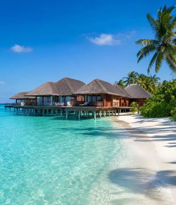 Maldives resort with overwater villas, turquoise lagoon, white sand beach, palm trees, and clear blue daytime sky.