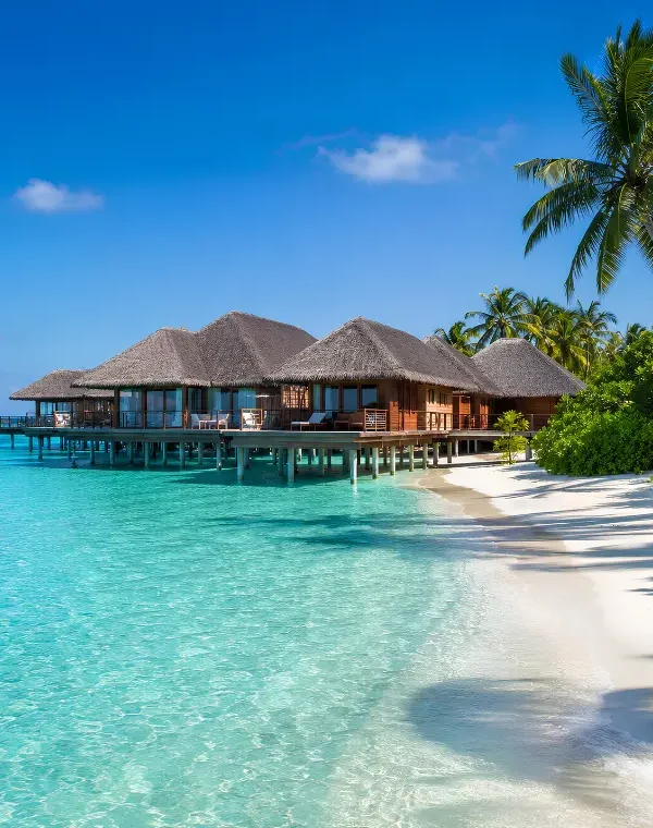 Maldives resort with overwater villas, turquoise lagoon, white sand beach, palm trees, and clear blue daytime sky.
