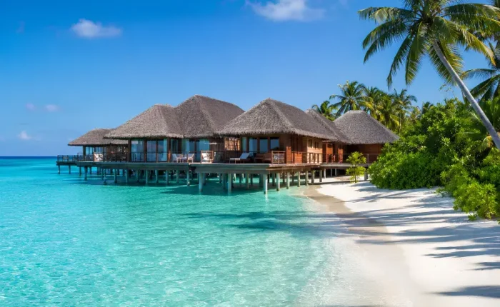 Maldives resort with overwater villas, turquoise lagoon, white sand beach, palm trees, and clear blue daytime sky.