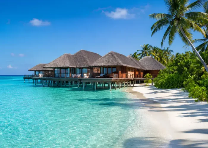 Maldives resort with overwater villas, turquoise lagoon, white sand beach, palm trees, and clear blue daytime sky.