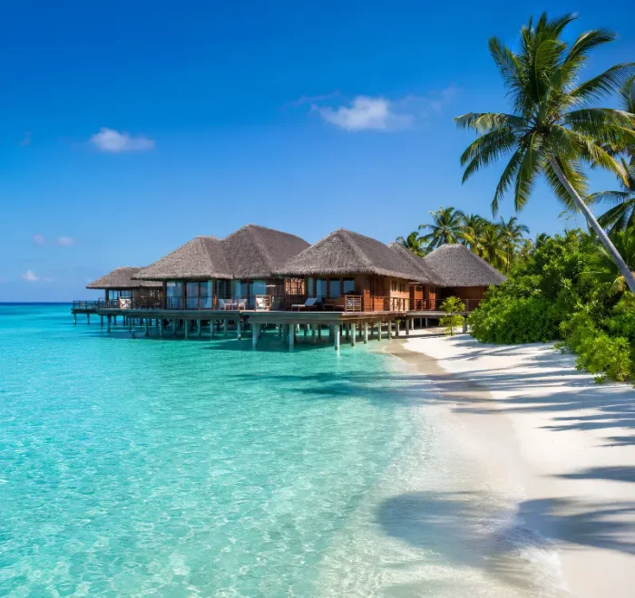 Maldives resort with overwater villas, turquoise lagoon, white sand beach, palm trees, and clear blue daytime sky.