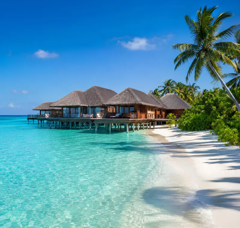 Maldives resort with overwater villas, turquoise lagoon, white sand beach, palm trees, and clear blue daytime sky.