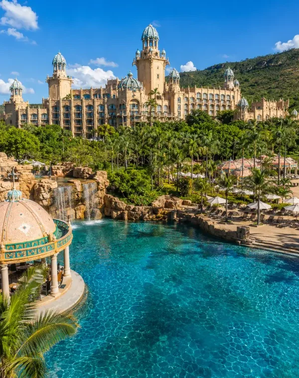 Luxury daytime view of Sun City South Africa with palace resort, turquoise pool, palm trees, and lush hills.