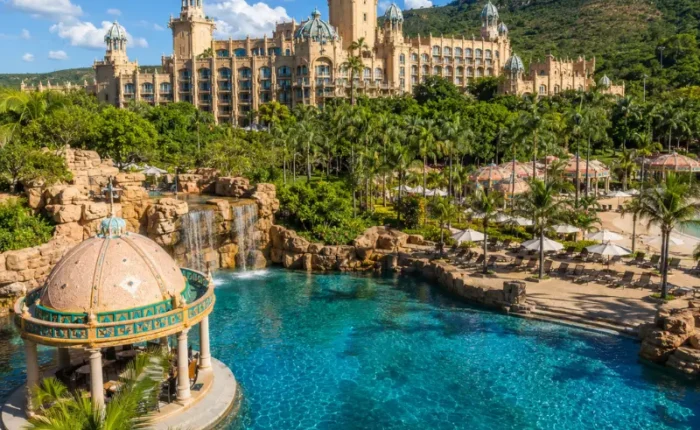 Luxury daytime view of Sun City South Africa with palace resort, turquoise pool, palm trees, and lush hills.