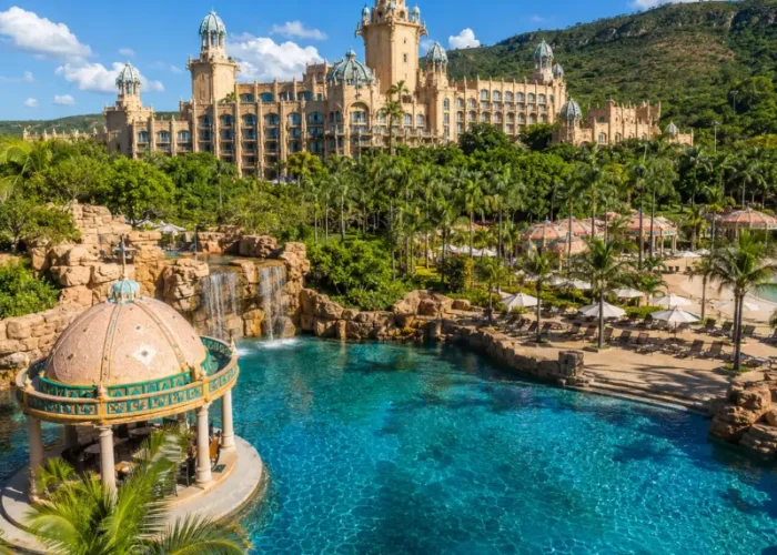 Luxury daytime view of Sun City South Africa with palace resort, turquoise pool, palm trees, and lush hills.