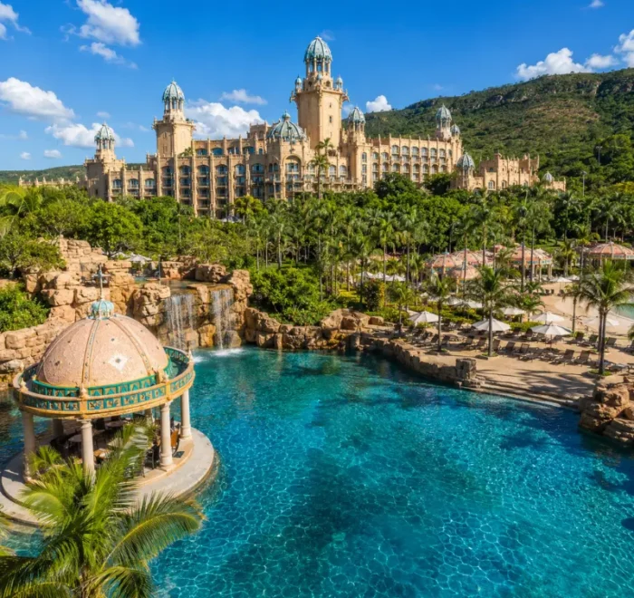 Luxury daytime view of Sun City South Africa with palace resort, turquoise pool, palm trees, and lush hills.