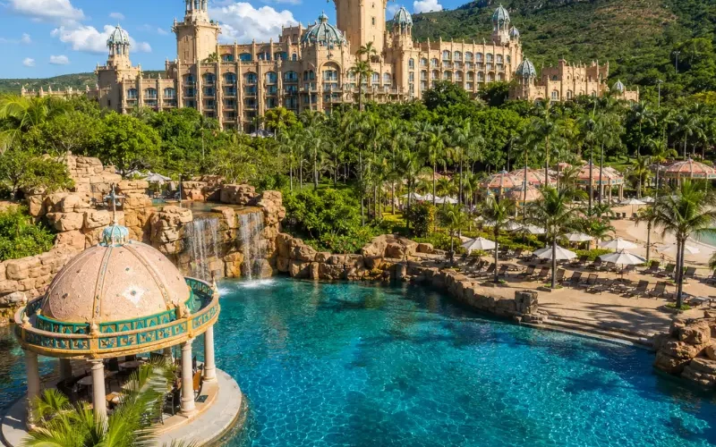 Luxury daytime view of Sun City South Africa with palace resort, turquoise pool, palm trees, and lush hills.
