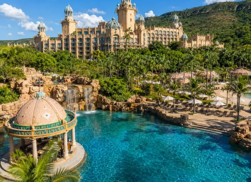 Luxury daytime view of Sun City South Africa with palace resort, turquoise pool, palm trees, and lush hills.