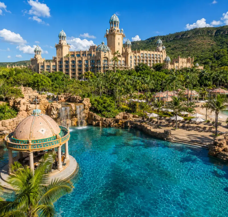 Luxury daytime view of Sun City South Africa with palace resort, turquoise pool, palm trees, and lush hills.