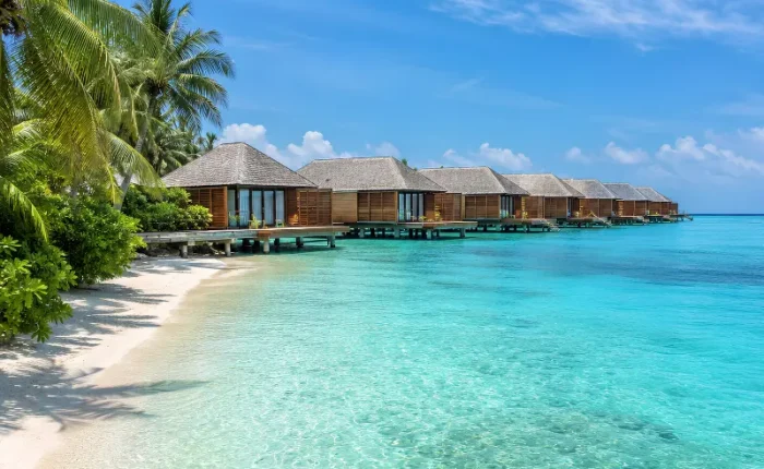 Maldives water villa resort with overwater bungalows, turquoise lagoon, white sand beach, and palm trees.