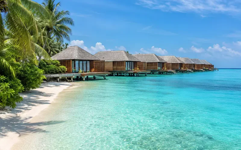 Maldives water villa resort with overwater bungalows, turquoise lagoon, white sand beach, and palm trees.