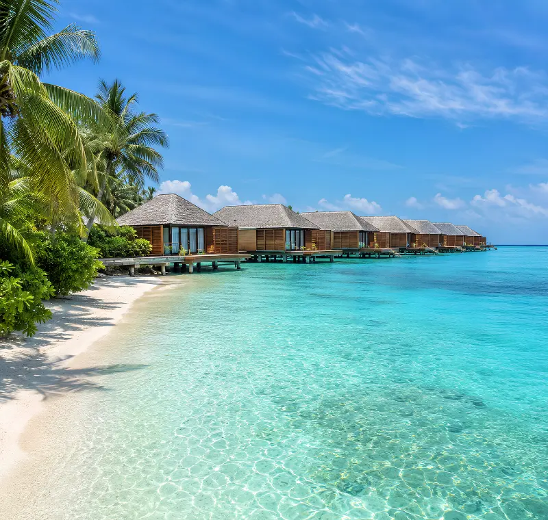 Maldives water villa resort with overwater bungalows, turquoise lagoon, white sand beach, and palm trees.