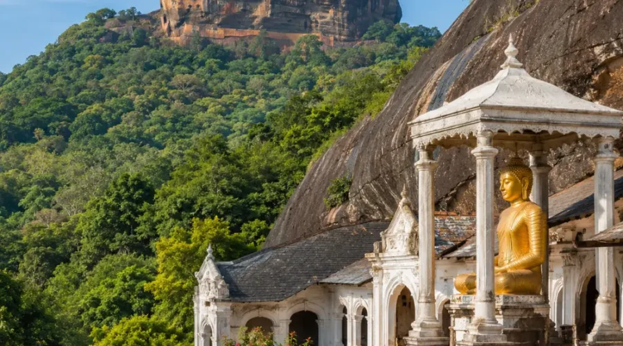 A breathtaking view of Sigiriya Rock Fortress rising dramatically above lush green forests alongside the ancient Dambulla Cave Temple with its golden Buddha statue, showcasing Sri Lanka's rich cultural heritage under a bright blue daytime sky.