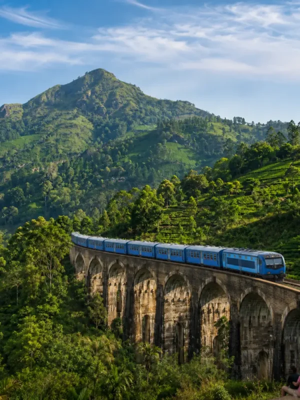 Nine Arches Bridge in Ella with a blue train, tourists enjoying the view, lush tea hills, and bright morning light.