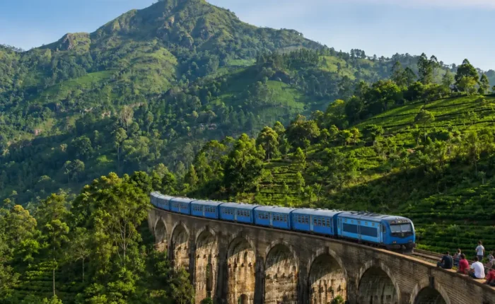 Nine Arches Bridge in Ella with a blue train, tourists enjoying the view, lush tea hills, and bright morning light.