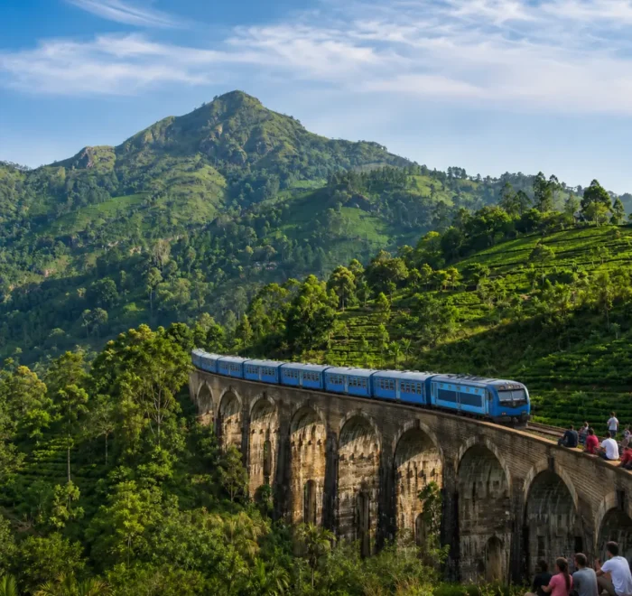 Nine Arches Bridge in Ella with a blue train, tourists enjoying the view, lush tea hills, and bright morning light.